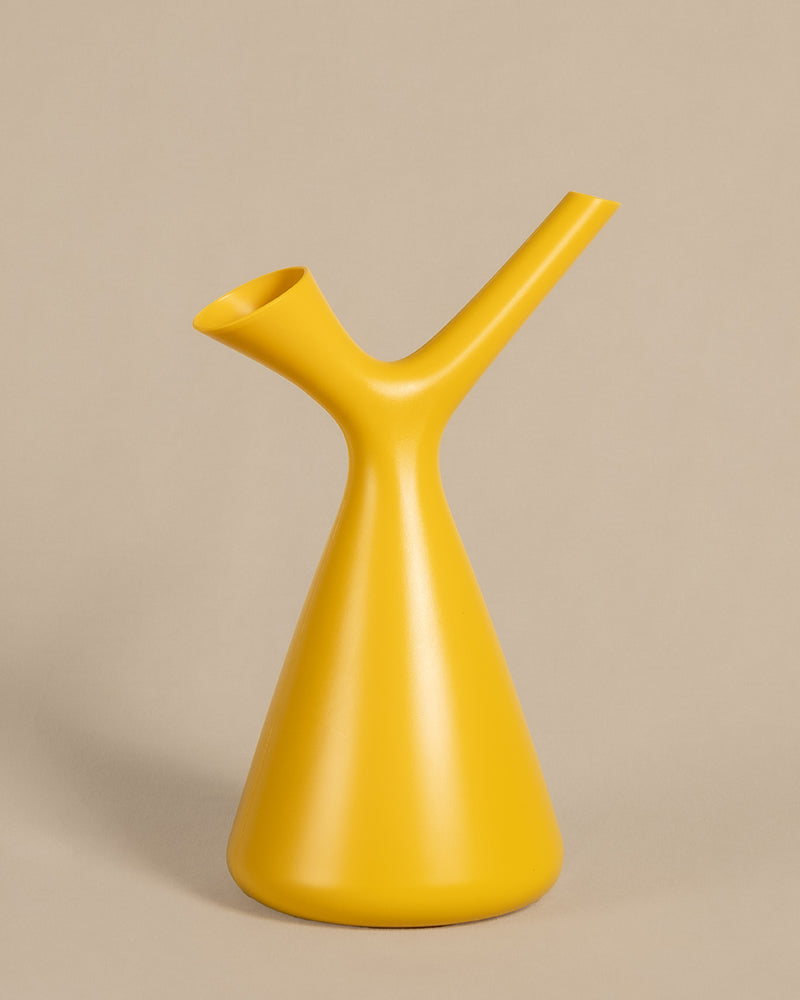 Eine moderne, minimalistische gelbe Gießkanne mit glatter, matter Oberfläche steht vor einem beigen Hintergrund. Die Plunge Gießkanne verfügt über ein einzigartiges, Y-förmiges Design mit einem Ausguss und einem Griff, die von einer konischen Basis abzweigen. Sie ist aus strapazierfähigem Polypropylen gefertigt und sowohl funktional als auch stilvoll.