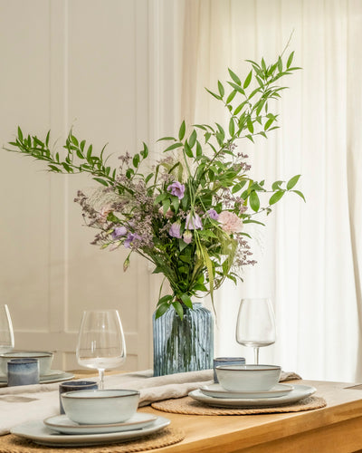 Ein mit hellblauem Geschirr, Weingläsern und einem geflochtenen Tischset gedeckter Esstisch wird elegant durch Purpurpoesie akzentuiert - ein hohes Blumenarrangement mit grünen Blättern und violetten Blüten in einer Glasvase. Weiße Vorhänge bilden den Hintergrund.