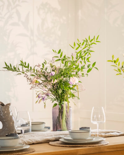 Die Vase Purpurpoesie, gefüllt mit gebundenen Blumen, grünem Laub und zarten Pastellblumen, steht auf einem gedeckten Esstisch mit Tellern, Schalen, Gläsern und Tischsets. Sanftes Licht wirft Schatten auf die Blätter dahinter - ein heiterer Mittelpunkt für jede Zusammenkunft.