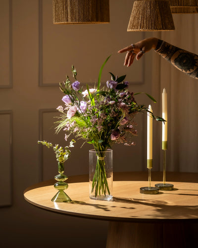 Eine tätowierte Hand greift nach "Purpurpoesie", einer Vase mit violetten und weißen Blumen auf einem runden Holztisch, neben zwei hohen Kerzen und einer kleinen Glas-Ode mit weißen Blüten. Warmes Licht unterstreicht die einladende Atmosphäre.