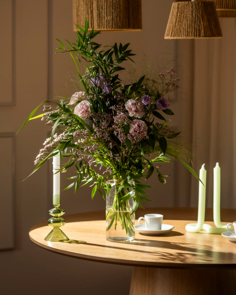 Das Purpurpoesie-Arrangement - eine hohe Glasvase, gefüllt mit violetten und rosafarbenen Blüten und Grünzeug - schmückt einen runden Holztisch mit grünen Kerzenhaltern, leuchtenden Kerzen und einer weißen Teetasse in sanftem, natürlichem Licht.