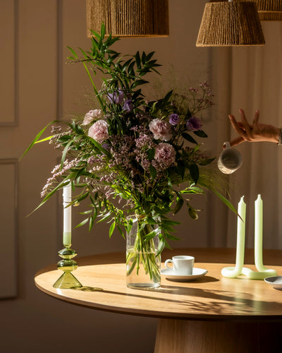 Die Purpurpoesie, ein üppiger Strauß lila und grüner Blüten in einer Glasvase, steht auf einem Holztisch mit grünen Kerzenhaltern, einer Teetasse und warmer Beleuchtung - eine Ode an die Gemütlichkeit von Anmut.