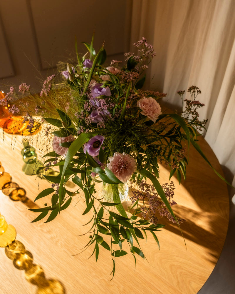 Der Purpurpoesie-Strauß aus rosa und violetten Blumen mit grünem Laub ruht auf einem hellen Holztisch im warmen Sonnenlicht, akzentuiert durch gelbe und grüne dekorative Glasperlen für einen schönen Touch.