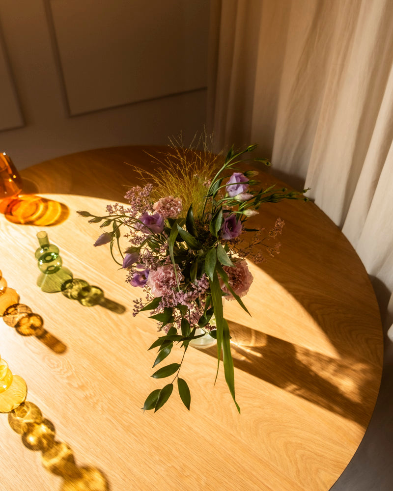 Der Purpurpoesie-Strauß in Lila und Rosa mit grünen Blättern ruht in einer Glasvase auf einem runden Holztisch neben buntem Glasdekor, das in Sonnenlicht getaucht ist - ein Fest der natürlichen Anmut.