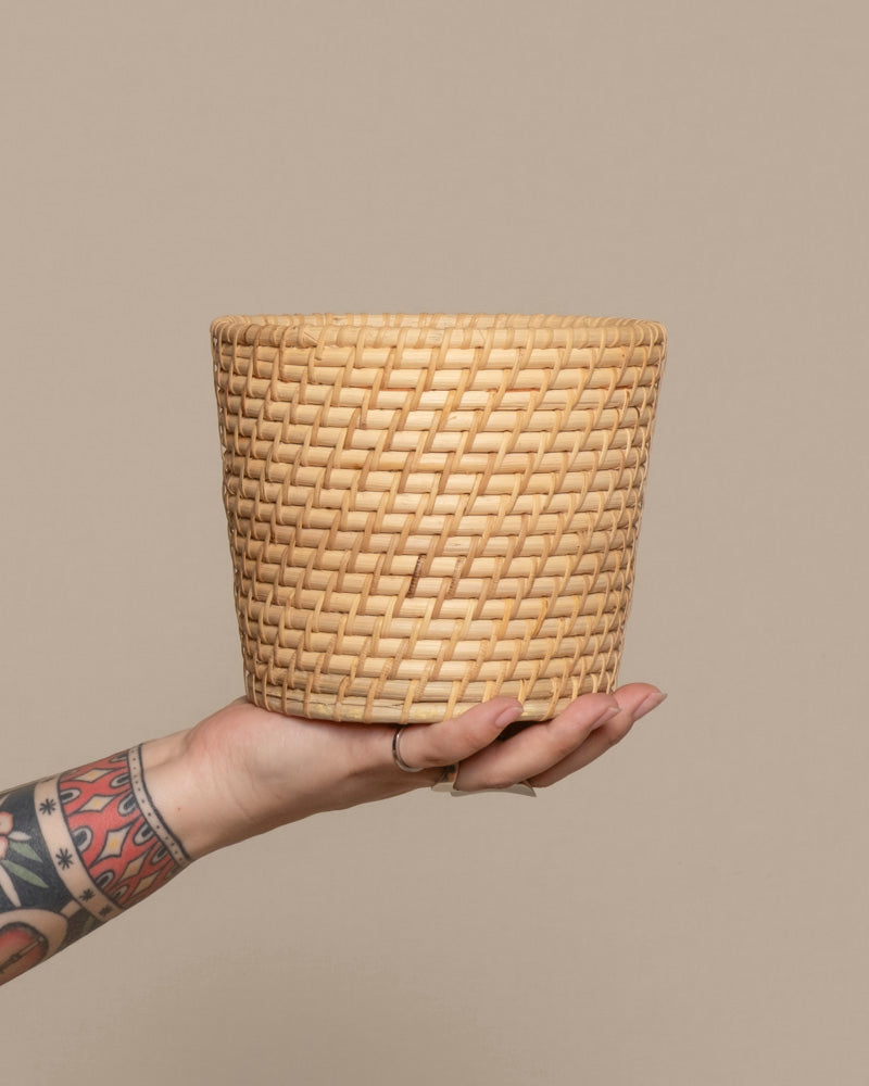 Eine tätowierte Hand hält den Pflanzenkorb aus Rattan (Calen | 14cm Ø) vor einem schlichten beigen Hintergrund hoch.