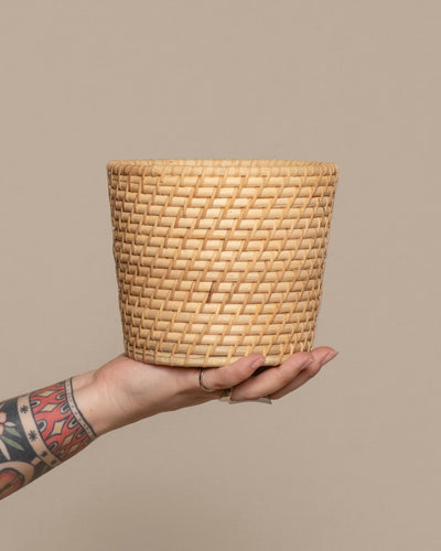 Eine tätowierte Hand hält den Pflanzenkorb aus Rattan (Calen | 14cm Ø) vor einem schlichten beigen Hintergrund hoch.