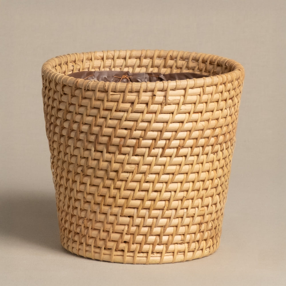 Der Pflanzenkorb aus Rattan (Calen | 16cm Ø) ist rund, hellbraun, handgefertigt und zeigt ein enges Flechtmuster. Er steht aufrecht vor einem schlichten beigen Hintergrund.
