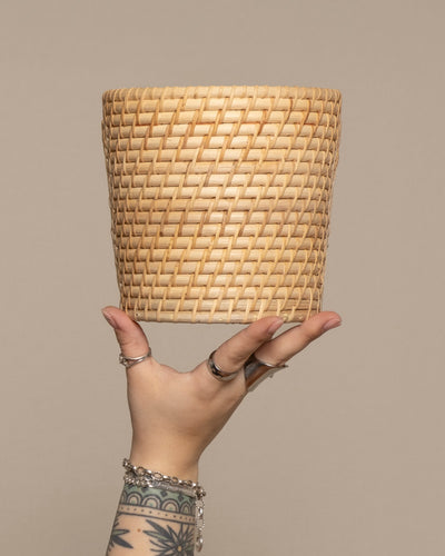 Eine tätowierte Hand mit Ringen und Armbändern hält den Pflanzenkorb aus Rattan (Calen | 16 cm Ø) hoch, einen stilvollen handgefertigten Rattankorb, der sich perfekt für Pflanzen eignet, vor einem schlichten beigen Hintergrund.