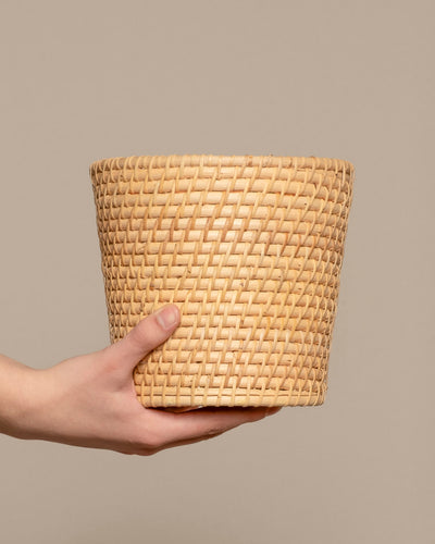Eine Hand hält den Pflanzenkorb aus Rattan (Calen | 18cm Ø) vor einem schlichten beigen Hintergrund.