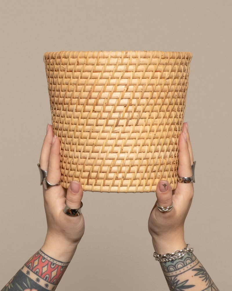 Zwei tätowierte Hände mit Ringen und Armbändern halten einen leeren, runden Pflanzenkorb aus Rattan (Calen | 22cm Ø) vor einem schlichten beigen Hintergrund.