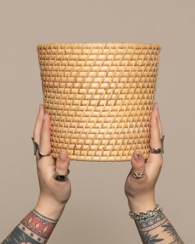 Zwei tätowierte Hände mit Ringen und Armbändern halten einen leeren, runden Pflanzenkorb aus Rattan (Calen | 22cm Ø) vor einem schlichten beigen Hintergrund.
