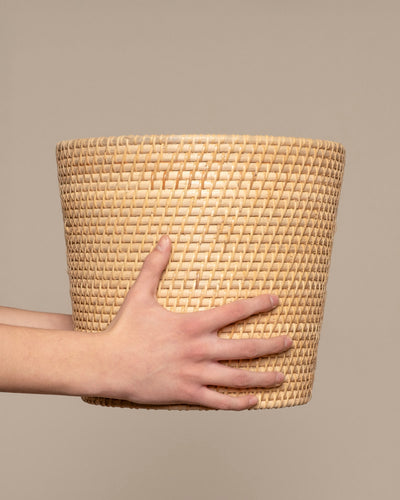 Eine Hand hält den Pflanzenkorb aus Rattan (Calen | 25cm Ø), einen großen, runden Rattankorb, vor einem schlichten beigen Hintergrund.