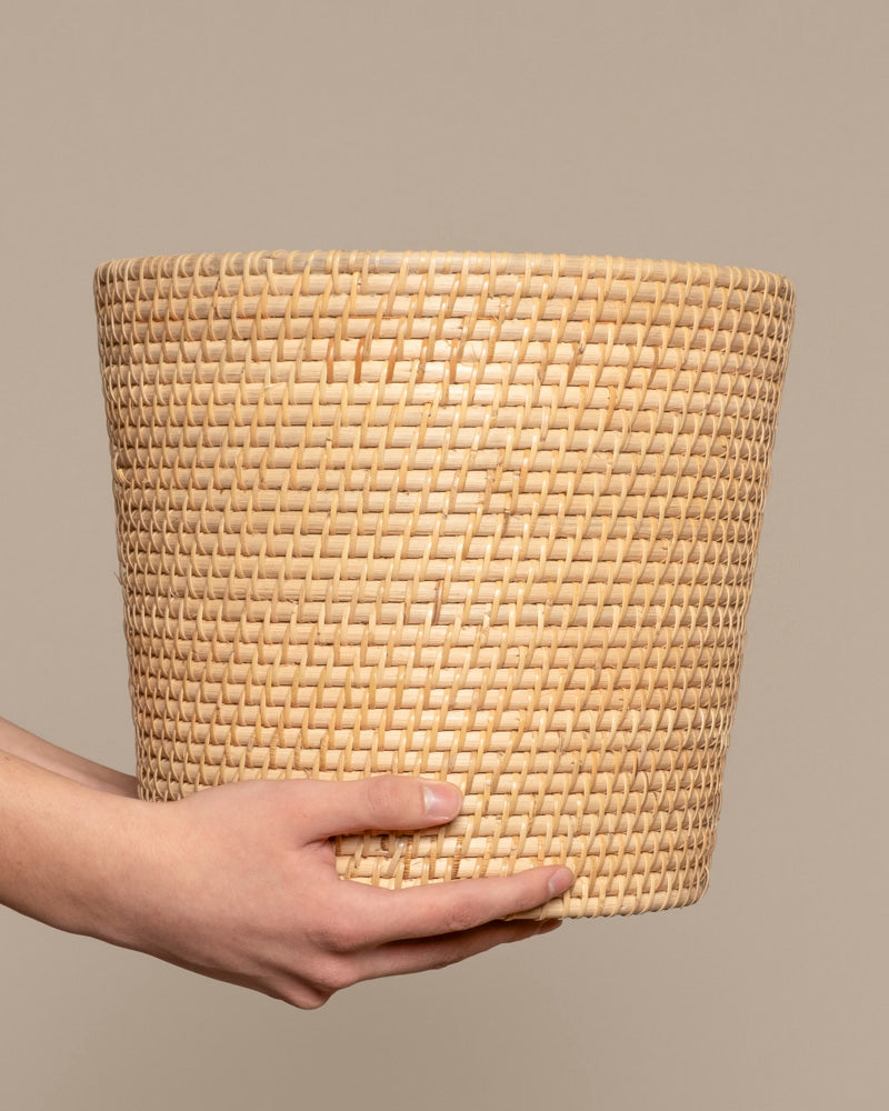 Eine Hand hält den Pflanzenkorb aus Rattan (Calen | 30cm Ø), einen großen, runden handgefertigten Rattankorb, vor einem schlichten beigen Hintergrund.