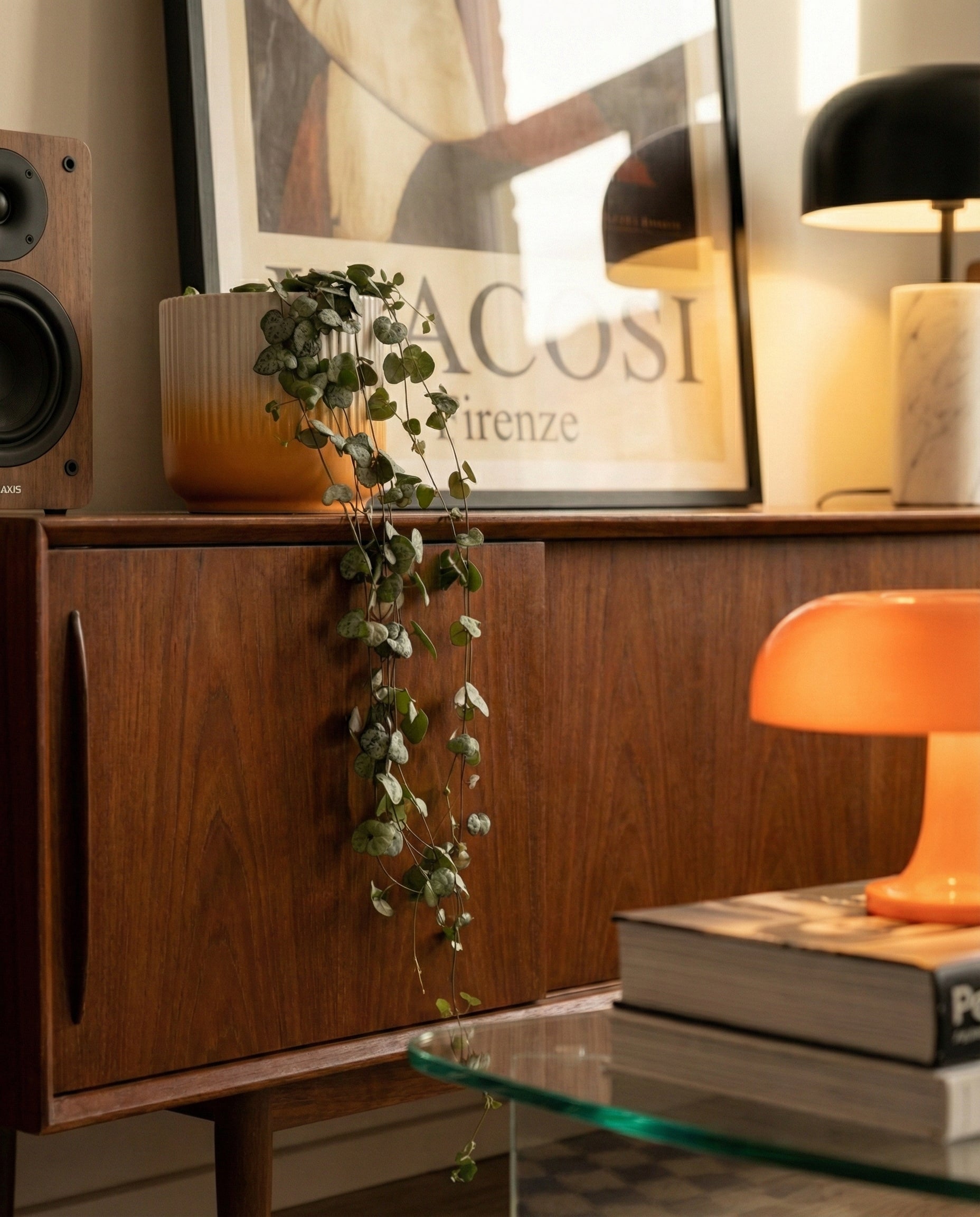 Ein modernes Holzsideboard aus der Mitte des Jahrhunderts enthält eine getopfte Leuchterpflanze, Bücher, einen Lautsprecher und zwei Lampen - eine schwarze und eine orangefarbene. Über der Anrichte lehnt ein gerahmtes Kunstwerk an der Wand.