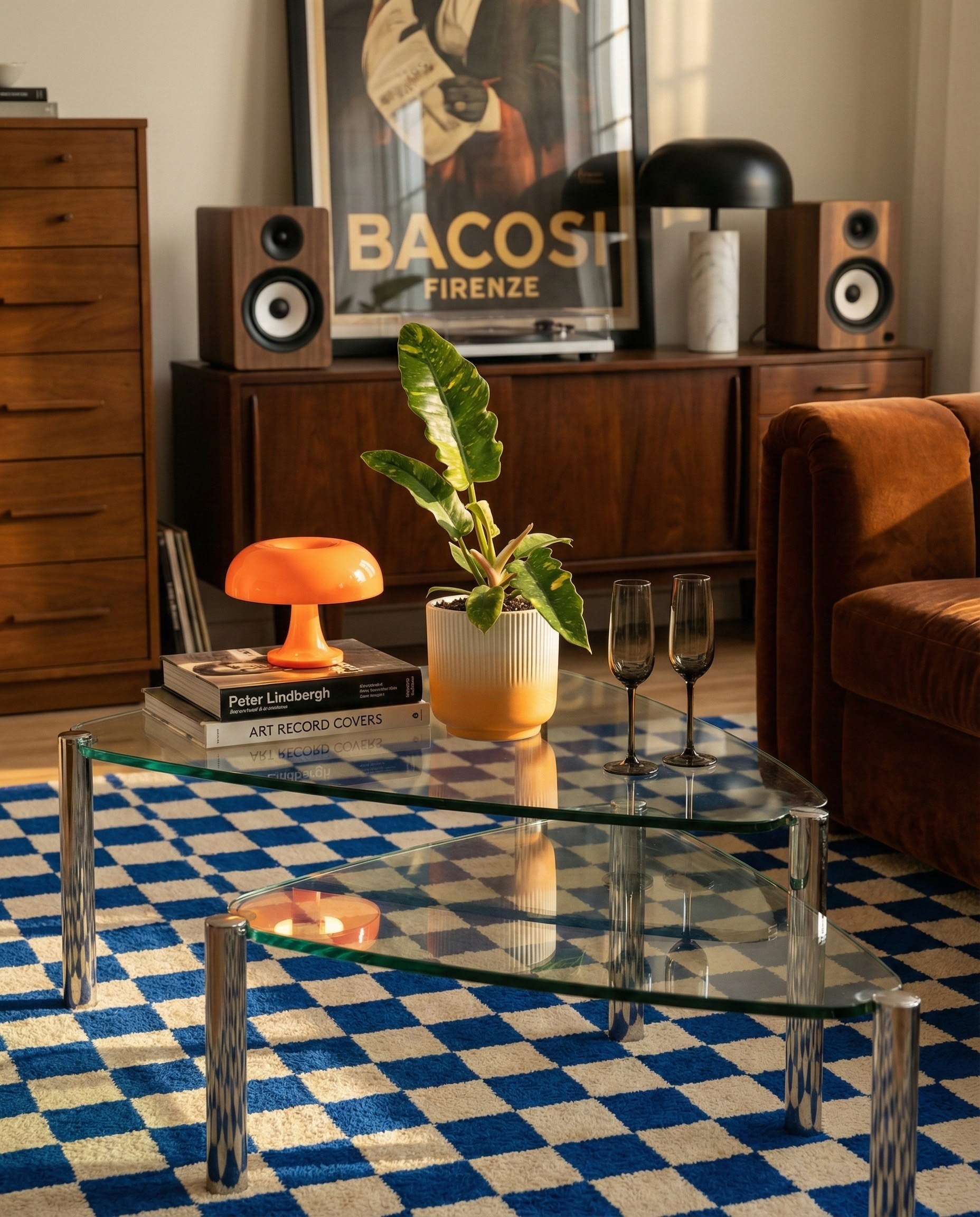 In einem stilvollen Wohnzimmer stehen ein gläserner Couchtisch mit Büchern, eine Topfpflanze im Keramik-Topf (Rilio | 14 cm ø) und zwei schwarze Gläser auf einem blau-karierten Teppich. Im Hintergrund stehen Lautsprecher, eine orangefarbene Retro-Lampe und ein 