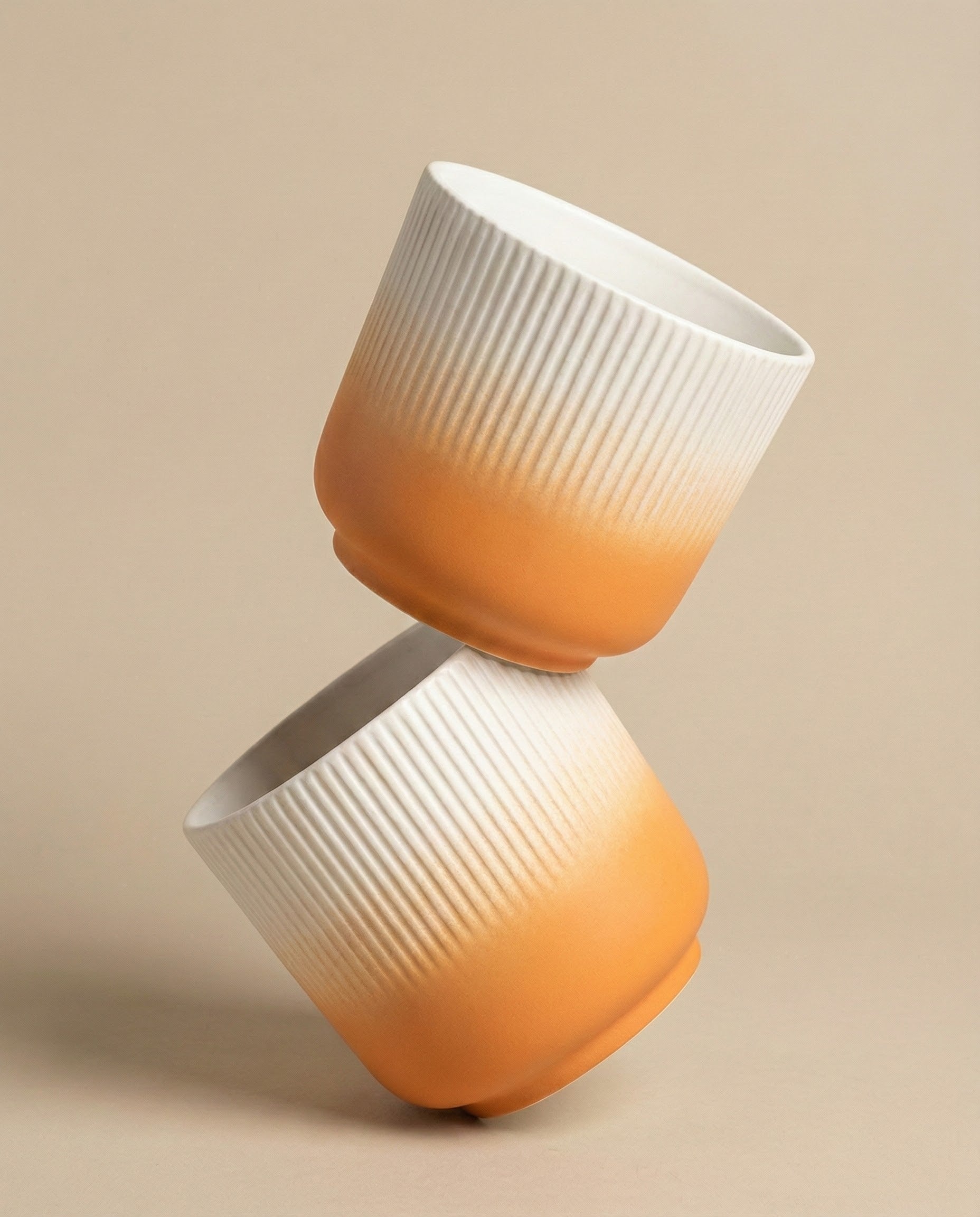 Zwei fey Keramik-Töpfe (Rilio | 14 cm ø) mit geriffelter Oberfläche und sanftem Farbverlauf von Weiß zu Orange sind gestapelt, wobei ein Topf auf dem anderen vor beigem Hintergrund balanciert.
