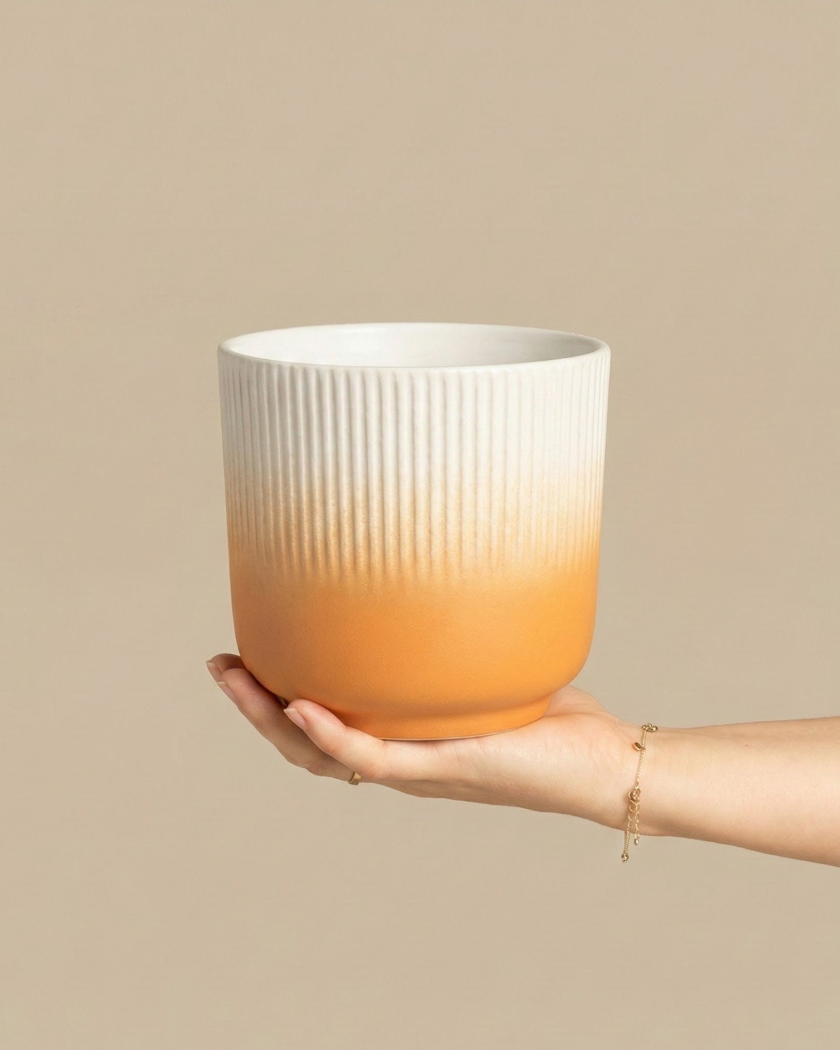 Eine Hand hält den Keramik-Topf (Rilio | 14 cm ø) mit einer strukturierten, gerippten weißen oberen Hälfte und einer glatten orangefarbenen unteren Hälfte vor einem beigen Hintergrund.