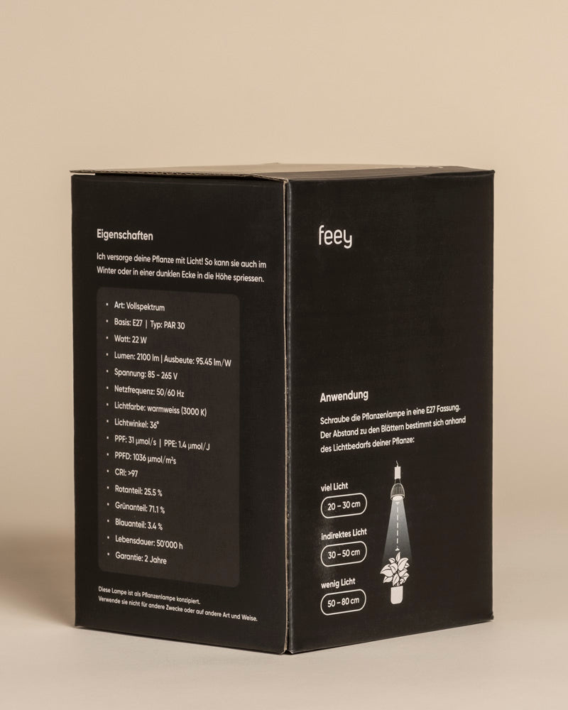 Ein schwarzer Feey-Produktkarton mit weißem deutschen Text enthält Angaben zu Spezifikationen und Gebrauchsanweisungen für die Pflanzenlampe 'Staila', einschließlich einer Abbildung des empfohlenen Lichtabstands für sonnenlicht-ähnliche Bedingungen bei Zimmerpflanzen.