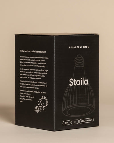 Eine schwarze Schachtel mit der Aufschrift Pflanzenlampe 'Staila' zeigt eine Strichzeichnung einer Glühbirne, einen deutschen Text und Symbole, die ihre Leistung von 22 W E27 hervorheben. Konzipiert für Zimmerpflanzen, bietet sie sonnenlicht-ähnliches Vollspektrum-Licht auf einem beigen Hintergrund.