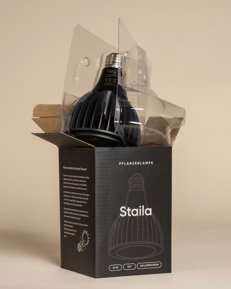 Die Pflanzenlampe 'Staila', eine schwarze LED-Lampe für Zimmerpflanzen, ist teilweise in klarem Kunststoff eingeschlossen und befindet sich in einer offenen schwarzen Schachtel, die mit Produktdetails und einer Strichzeichnung der Lampe auf beigem Hintergrund beschriftet ist.