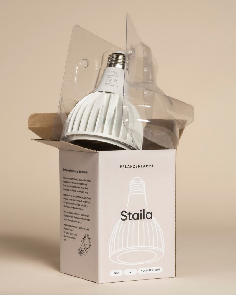 Eine weiße Pflanzenlampe 'Staila', teilweise aus durchsichtigem Plastik, sitzt in einer offenen Schachtel mit der Aufschrift "Staila" mit einer Lampenillustration und Details, ideal für Zimmerpflanzen, vor einem beigen Hintergrund.
