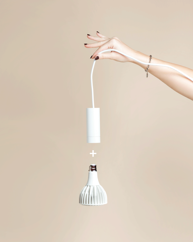 Eine Hand mit lackierten Nägeln hält eine weiße Pendelleuchte 'Corda' über einer passenden weißen LED-Glühbirne, die mit einem Pluszeichen dazwischen abgebildet ist - perfekt für eine stilvolle Beleuchtung von Zimmerpflanzen vor einem beigen Hintergrund.