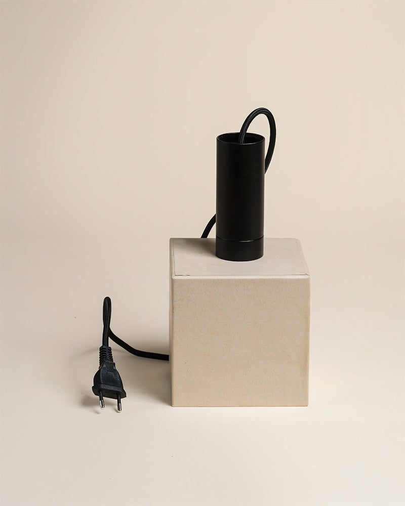 Eine schwarze zylindrische Pendelleuchte 'Corda' mit Kordel und Stecker sitzt auf einem beigen Würfel vor einem hellen Hintergrund. Die Kordel hängt an der Pendelleuchte, und der Stecker liegt neben dem Würfel.