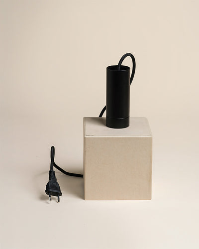 Eine schwarze zylindrische Pendelleuchte 'Corda' mit Kordel und Stecker sitzt auf einem beigen Würfel vor einem hellen Hintergrund. Die Kordel hängt an der Pendelleuchte, und der Stecker liegt neben dem Würfel.