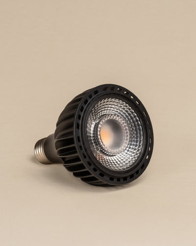 Die Pflanzenlampe 'Staila' in Schwarz mit metallischem Sockel und geriffeltem Gehäuse ruht seitlich auf einem einfarbig beigen Hintergrund. Ihre reflektierende Linse ist sichtbar - perfekt, um Sonnenlicht auf Ihre Zimmerpflanzen zu bringen.