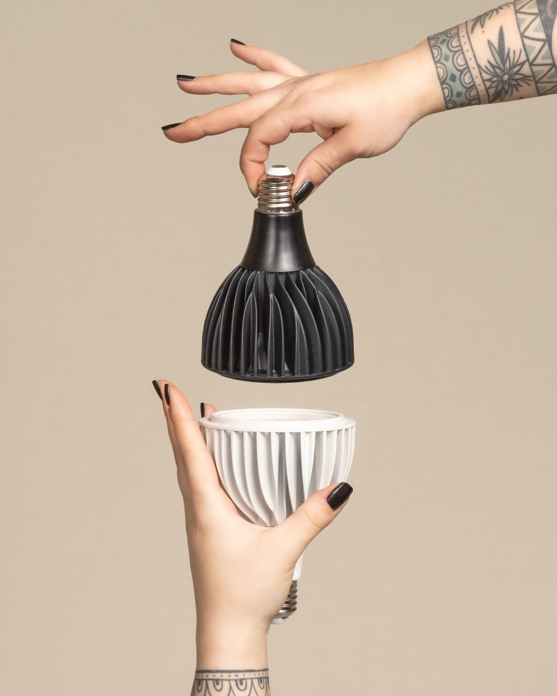 Zwei tätowierte Hände mit schwarzem Nagellack halten die Pflanzlampe 'Staila' - eine schwarze und eine weiße geometrische LED-Glühbirne - vor einem beigen Hintergrund, wobei die Glühbirnen so ausgerichtet sind, als ob sie verbunden werden sollen.