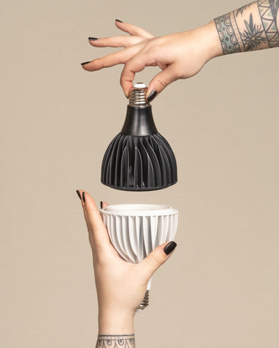 Zwei tätowierte Hände mit schwarzem Nagellack halten die Pflanzlampe 'Staila' - eine schwarze und eine weiße geometrische LED-Glühbirne - vor einem beigen Hintergrund, wobei die Glühbirnen so ausgerichtet sind, als ob sie verbunden werden sollen.