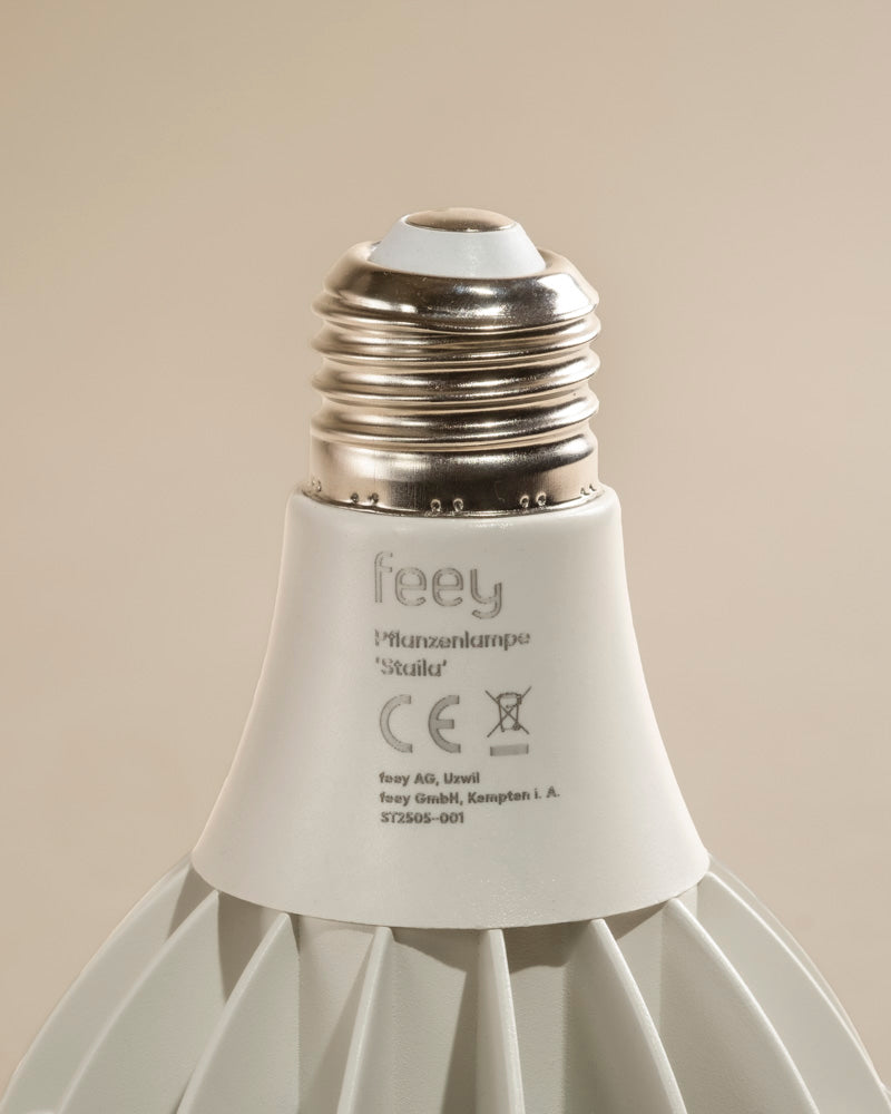 Nahaufnahme der weißen Pflanzenlampe 'Staila' mit metallischem Schraubsockel, mit Marken- und Zertifizierungsetiketten auf dem Korpus vor einem schlichten beigen Hintergrund - perfekt für sonnenlichtliebende Zimmerpflanzen.