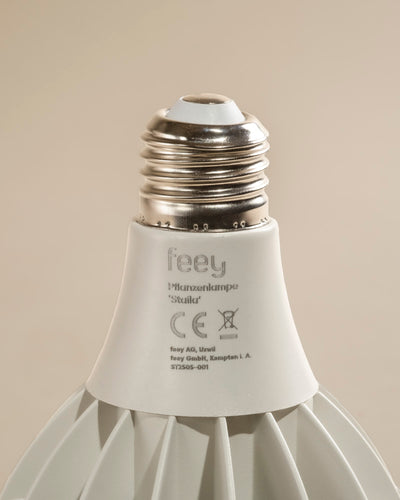 Nahaufnahme der weißen Pflanzenlampe 'Staila' mit metallischem Schraubsockel, mit Marken- und Zertifizierungsetiketten auf dem Korpus vor einem schlichten beigen Hintergrund - perfekt für sonnenlichtliebende Zimmerpflanzen.