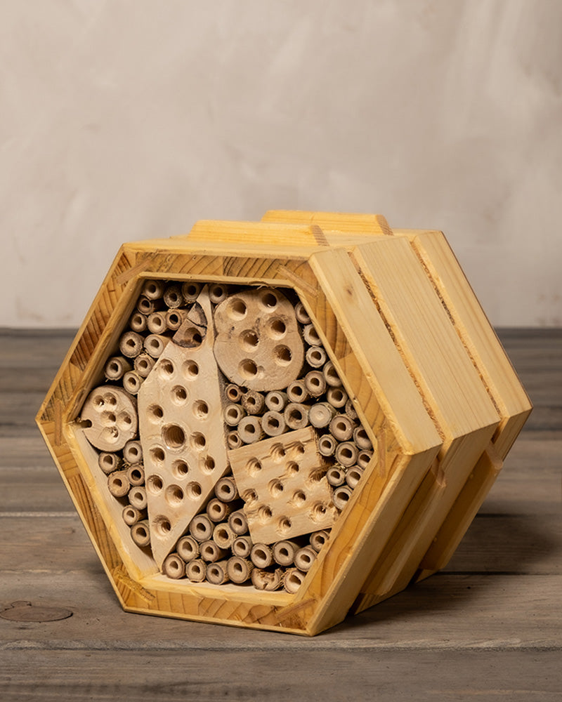Bienenhotel aus Bambus, mit Röhren in verschiedenen Größen und gebohrten Löchern, das so konzipiert ist, dass es einzelgängerische Wildbienen anlockt und ihnen einen Nistplatz bietet. Der Hintergrund ist eine neutrale Holzoberfläche und eine hellgraue Wand.