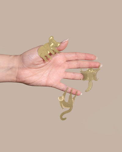 Eine Hand mit langen Nägeln hält drei goldene Ausschnitte in Tierform an ihren Fingern. Von links nach rechts ähneln die Formen einem Koala, einem Elefanten und einem Affen. Der Hintergrund ist in einem kräftigen, hellen Beige gehalten – ein charmanter Akzent für jeden Raum.