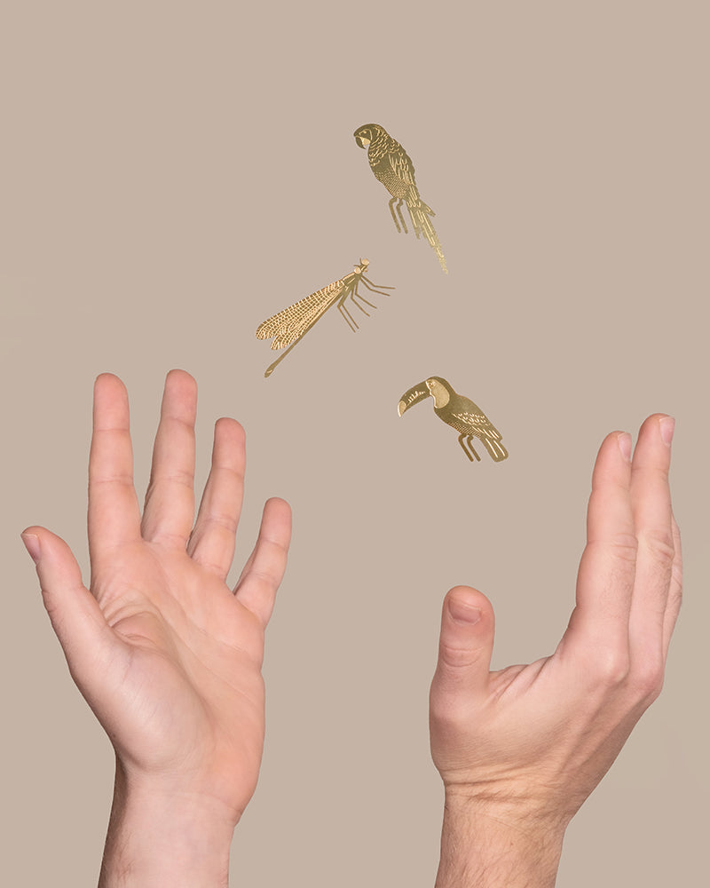 Vor einem beigen Hintergrund sind zwei offene Hände platziert, zwischen denen drei goldene, illustrierte Insekten und Vögel schweben. Die Hände scheinen den Tierischen Pflanzenhänger sanft loszulassen oder aufzufangen, was der Szene eine skurrile, magische Atmosphäre verleiht.