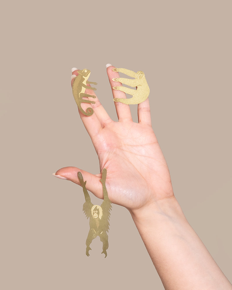 Eine Hand mit ausgebreiteten Fingern vor beigem Hintergrund. Auf jeder Fingerspitze sind dekorative Tierchen zu sehen: ein goldfarbenes Chamäleon auf dem Zeigefinger, ein roter Panda auf dem Mittelfinger, ein Orang-Utan, der am kleinen Finger hängt, und ein leerer Ringfinger, der auf einen Tierischen Pflanzenhänger wartet.
