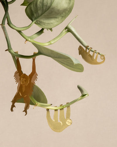 Zwei verzierte Lesezeichen aus Metall, eines in Form eines Orang-Utans und das andere in Form eines Faultiers, hängen an den Zweigen einer grünen Pflanze und ergänzen Ihren Tierischen Pflanzenhänger. Der Hintergrund ist schlicht beige.