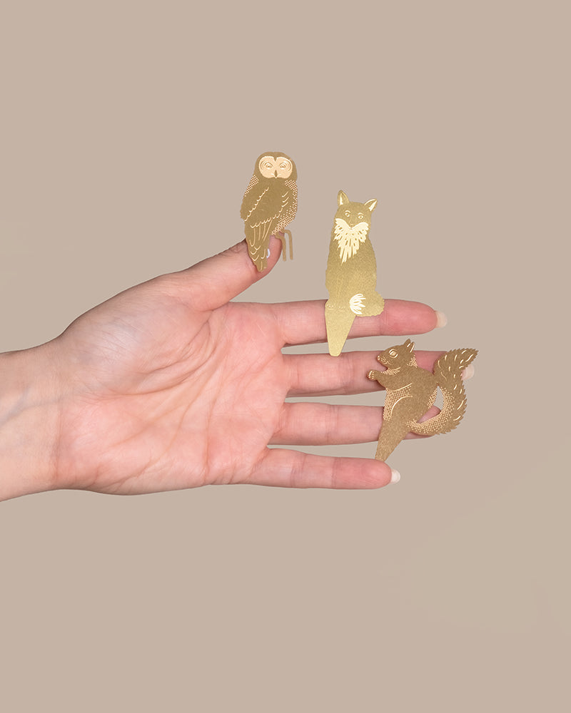 Eine Hand hält drei goldene Tierische Pflanzenhänger vor einem beigen Hintergrund. Diese dekorativen Tierchen stellen eine Eule, einen Fuchs und ein Eichhörnchen dar, alle mit komplizierten, stilisierten Details gestaltet. Die Hand ist geöffnet, die Nadeln ruhen auf den Fingern und der Handfläche.