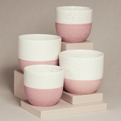 Vier handhergestellte Keramiktassen sind auf beigen Blöcken angeordnet. Die Tassen sind in der oberen Hälfte weiß und in der unteren Hälfte hellrosa, mit einer gesprenkelten Textur. Sie variieren in der Größe und werden vor einem neutralen Hintergrund präsentiert, perfekt für das Keramik-Topfset 'Variado' (2 × 16, 2 × 14).