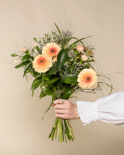 Eine Hand in einem weißen Ärmel hält den Überraschungs-Blumenstrauß - helle pfirsichfarbene Gerbera-Gänseblümchen, kleine Rosen und Grünzeug - ideal als Geschenk oder entzückende Überraschung, vor einem schlichten beigen Hintergrund.