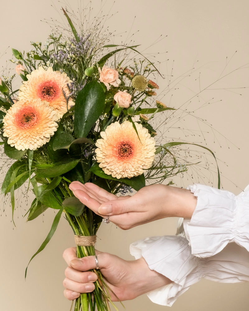 Eine Person in einem weißen, gerüschten Ärmel hält einen Überraschungs-Blumenstrauß aus pfirsichfarbenen Gerbera-Gänseblümchen, kleinen Rosen und Grünzeug - ein elegantes Geschenk vor einem neutralen Hintergrund.