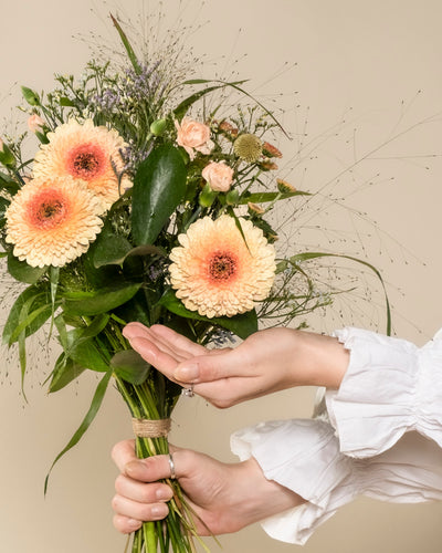 Eine Person in einem weißen, gerüschten Ärmel hält einen Überraschungs-Blumenstrauß aus pfirsichfarbenen Gerbera-Gänseblümchen, kleinen Rosen und Grünzeug - ein elegantes Geschenk vor einem neutralen Hintergrund.