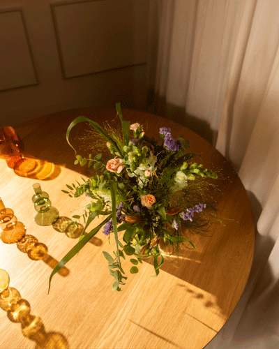 Der Überraschungs-Blumenstrauß mit bunten Blumen und Grün in einer Vase steht auf einem runden Holztisch neben Glas-Deko-Sonnenlicht fällt hinein. So schenken Sie überraschende Blumen als liebevolle Aufmerksamkeit.
