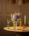 Der Überraschungs-Blumenstrauß mit einer violetten Hortensie und getrockneten Gräsern steht auf einem runden Tisch, umgeben von farbenfrohen Glas-Kerzenhaltern und einer brennenden Spitzkerze - ideal zum Verschenken von Blumen oder als charmante Überraschung bei warmem Licht.