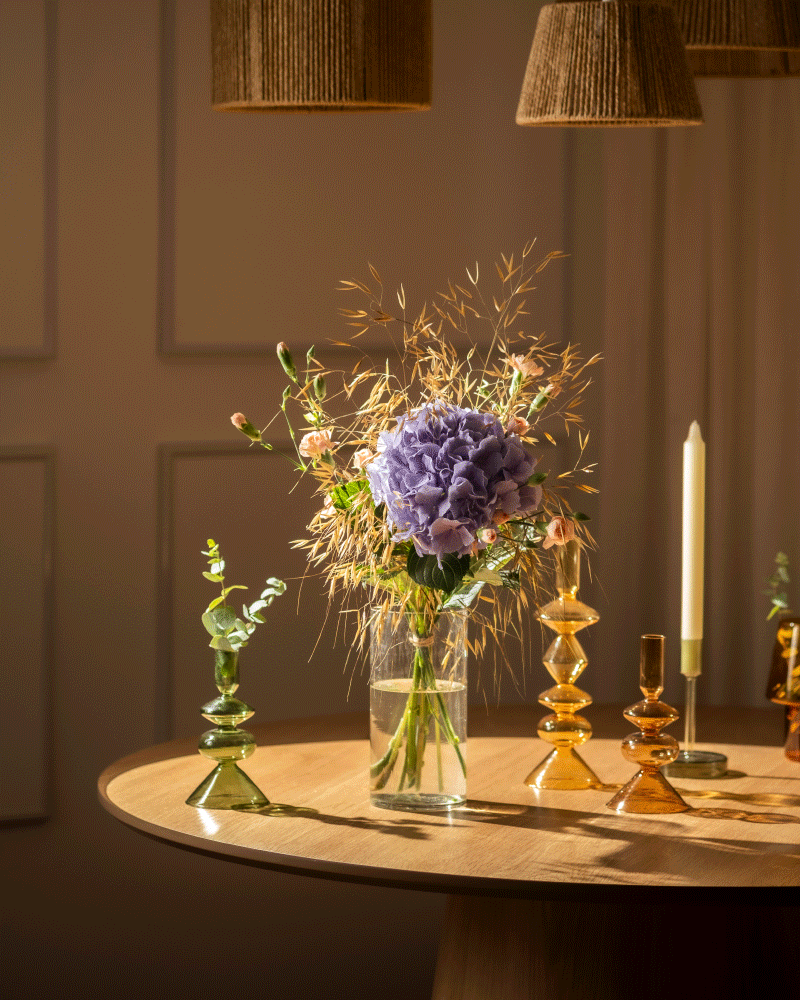 Der Überraschungs-Blumenstrauß mit einer violetten Hortensie und getrockneten Gräsern steht auf einem runden Tisch, umgeben von farbenfrohen Glas-Kerzenhaltern und einer brennenden Spitzkerze - ideal zum Verschenken von Blumen oder als charmante Überraschung bei warmem Licht.