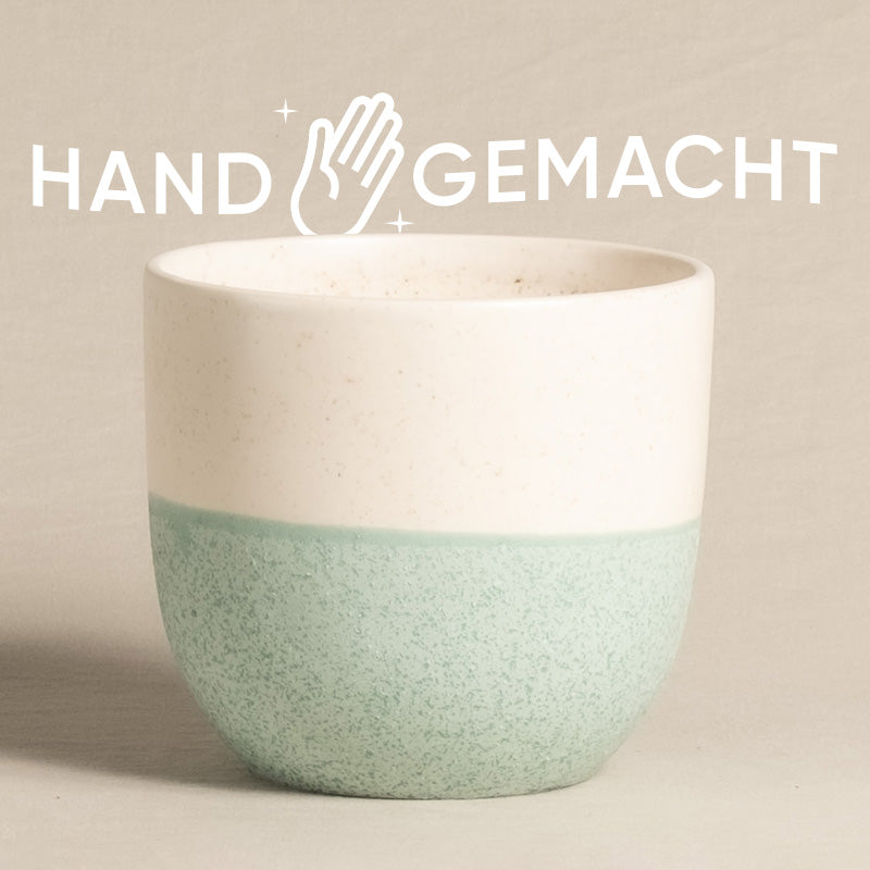 Eine Keramiktasse mit einer glatten weißen oberen Hälfte und einer strukturierten mintgrünen unteren Hälfte steht zentriert vor einem beigen Hintergrund. Über der Tasse sind die Worte „HAND GEMACHT“ und ein stilisiertes Handsymbol in Weiß zu sehen, was die handwerkliche Qualität unserer Kollektion feey Keramik-Topf (Variado | 14 cm ⌀) widerspiegelt.