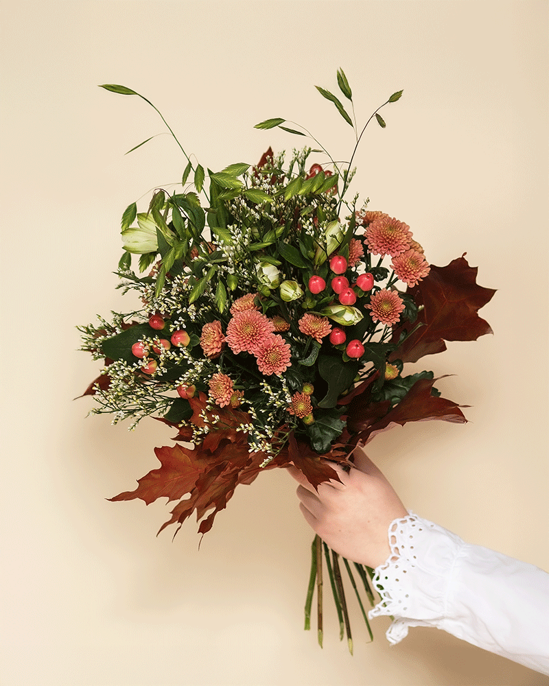 Eine Hand in einer weißen Spitzenmanschette präsentiert den Herbstfreude-Strauß - rote Beeren, rosafarbene Chrysanthemen, grüne Blätter, weiße Füllblumen und braunes Laub - und schafft damit ein fröhliches Farbenspiel vor einem beigen Hintergrund.