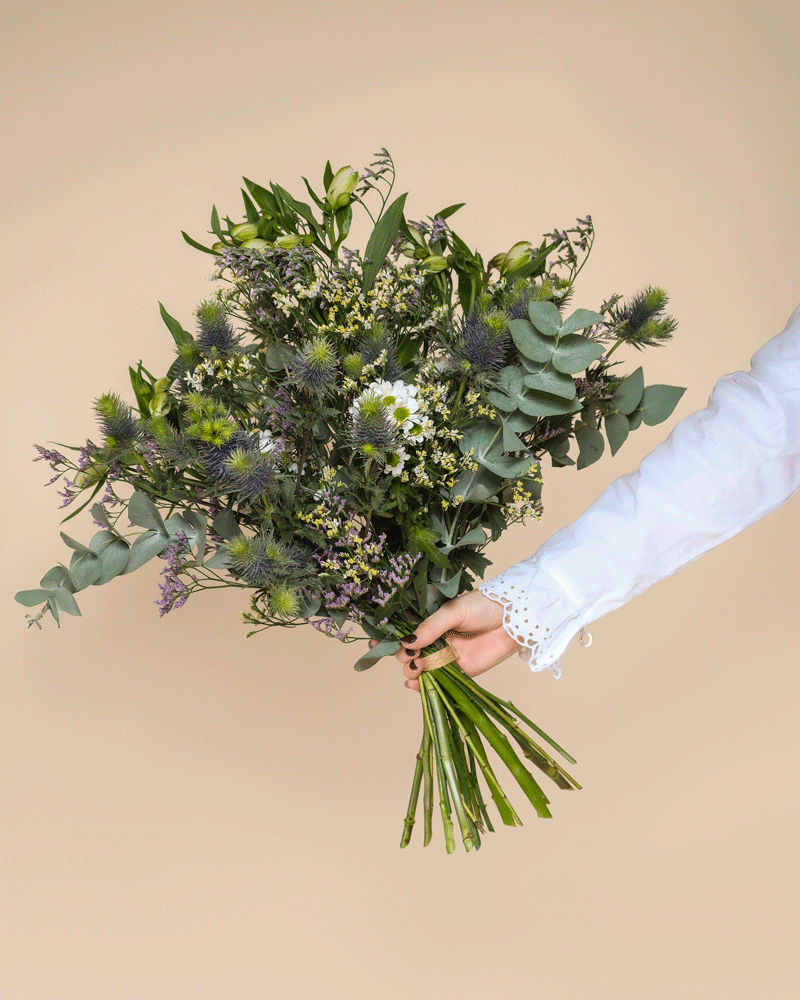 Eine Person im weißen Langarmhemd hält den Blumenstrauß "Nebelmeer" mit zarten weißen und lila Blüten sowie Morgentau vor einem schlichten beigen Hintergrund.