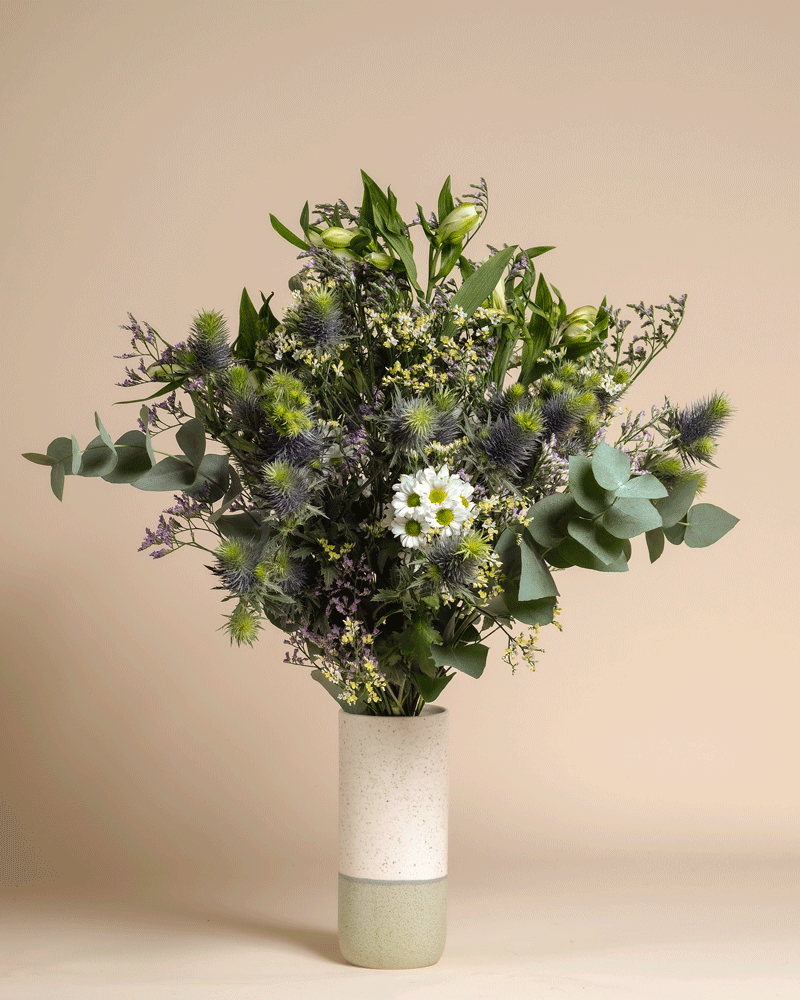 Der Blumenstrauß Nebelmeer vereint weiße Gänseblümchen, violette Disteln, Eukalyptus und gelbe Blüten in einer schlichten zweifarbigen Keramikvase vor beigem Hintergrund.